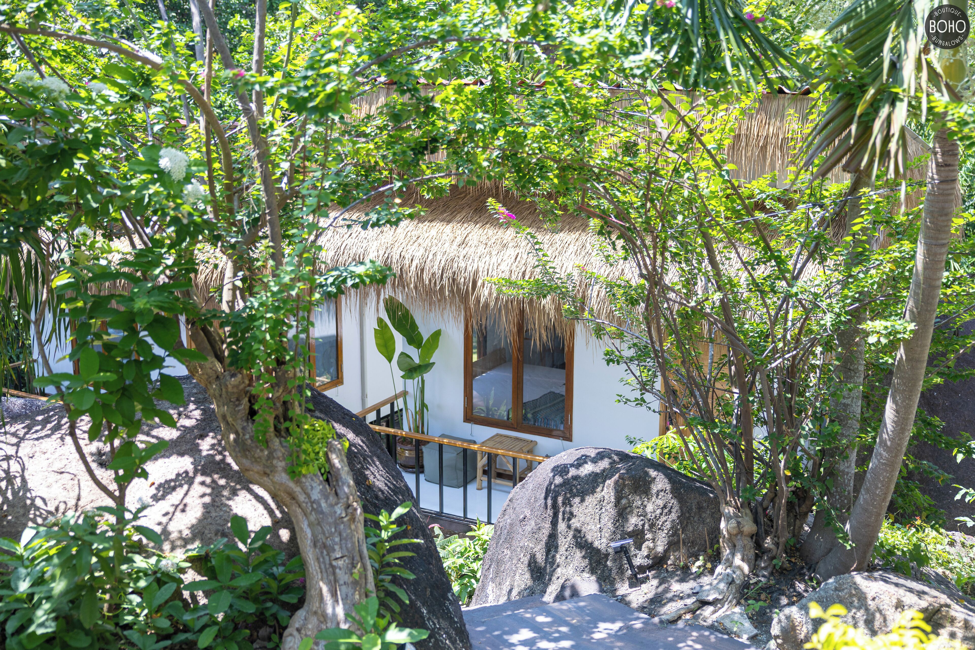 A thatched-roof house with a balcony, surrounded by lush greenery and large rocks.