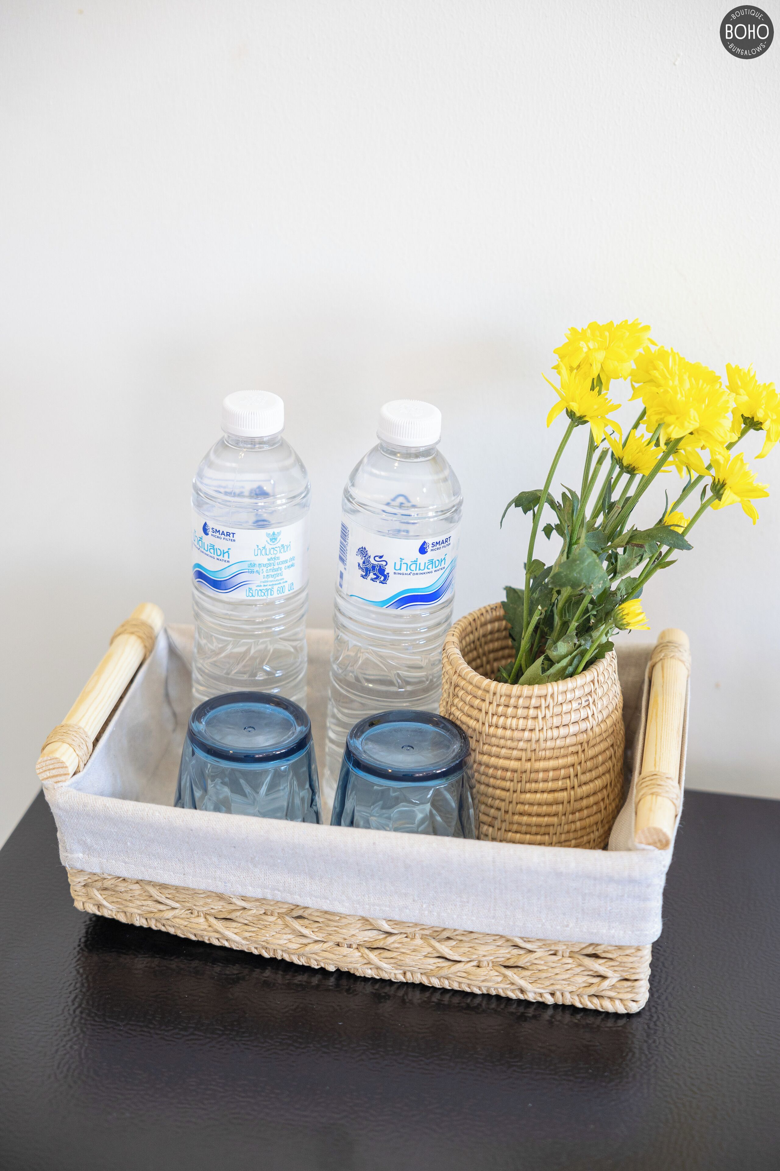 Una bandeja de mimbre con dos botellas de agua, un florero con flores amarillas y dos recipientes de vidrio sobre una superficie oscura.