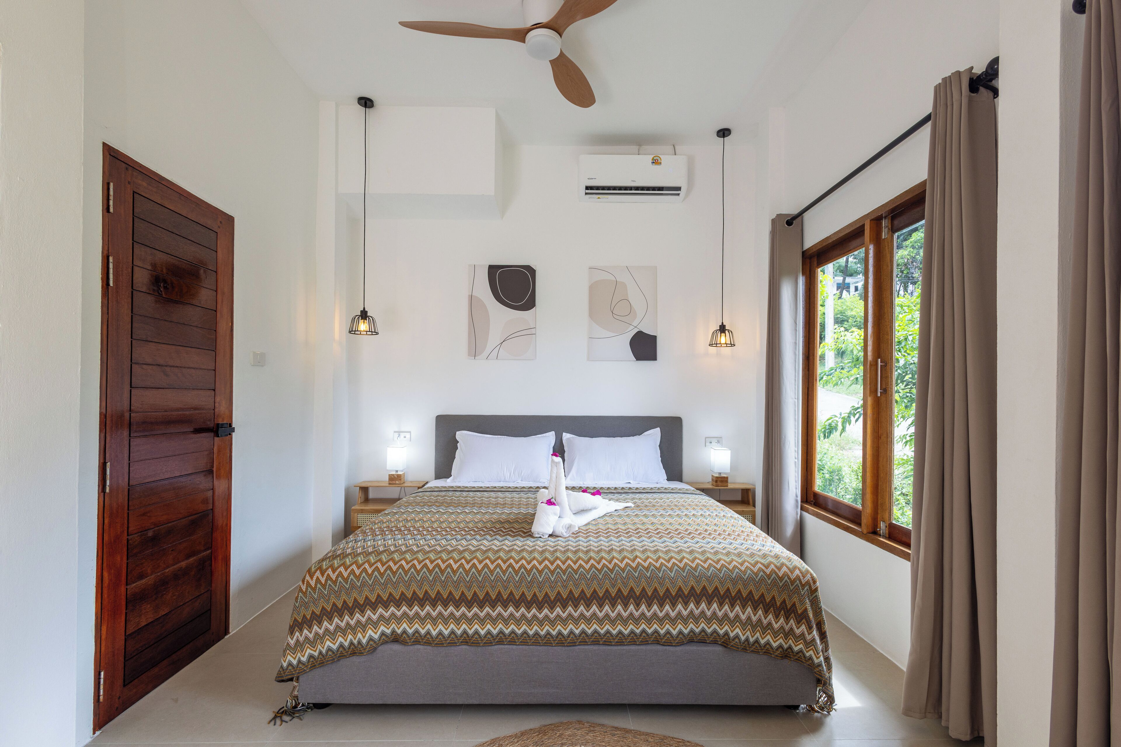 A bedroom with a wooden door, a ceiling fan, an air conditioning unit, a bed with a patterned bedspread, two bedside lamps, and a window with curtains.