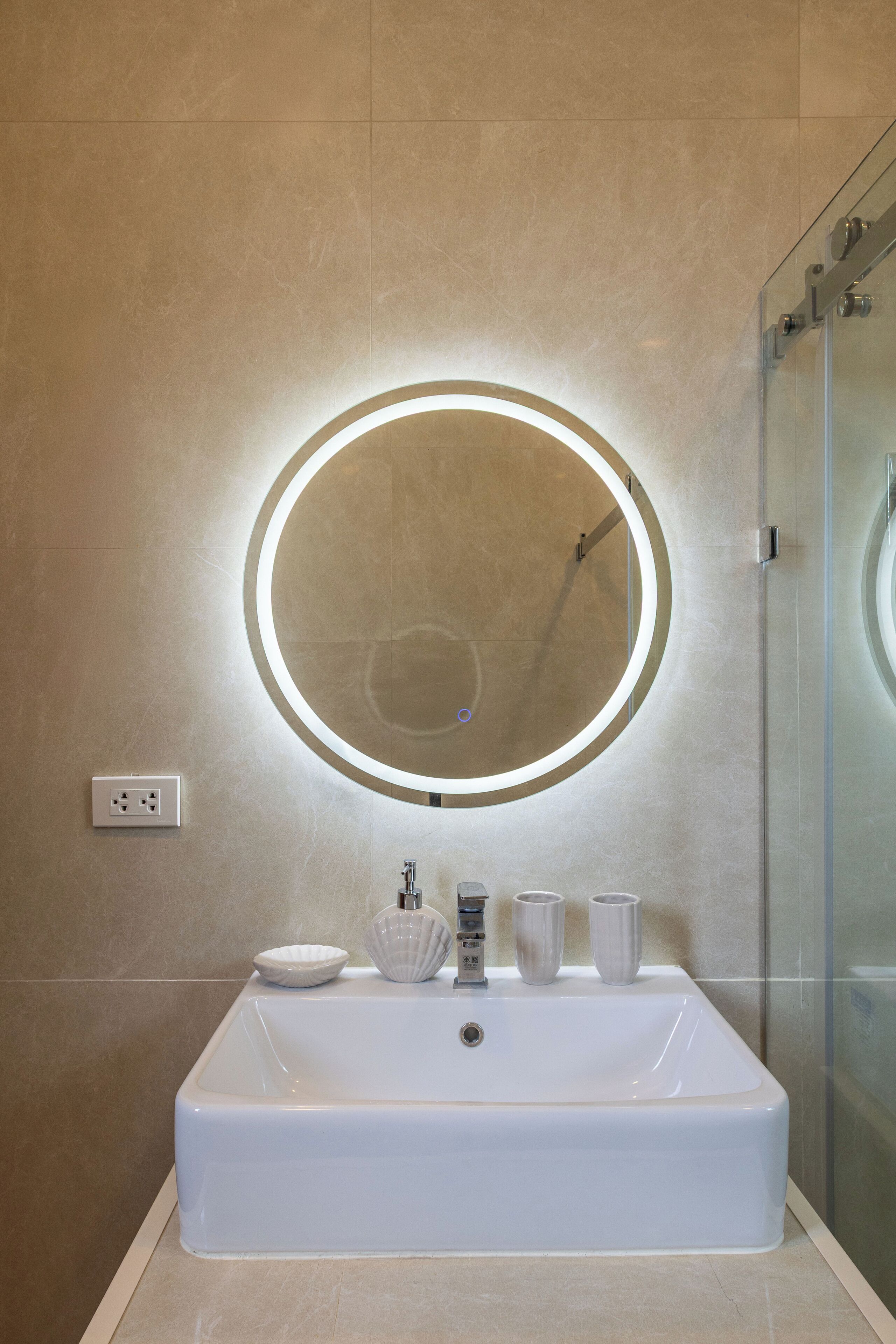 A modern bathroom with a large rectangular sink, a round mirror with a light, and a glass shower enclosure.