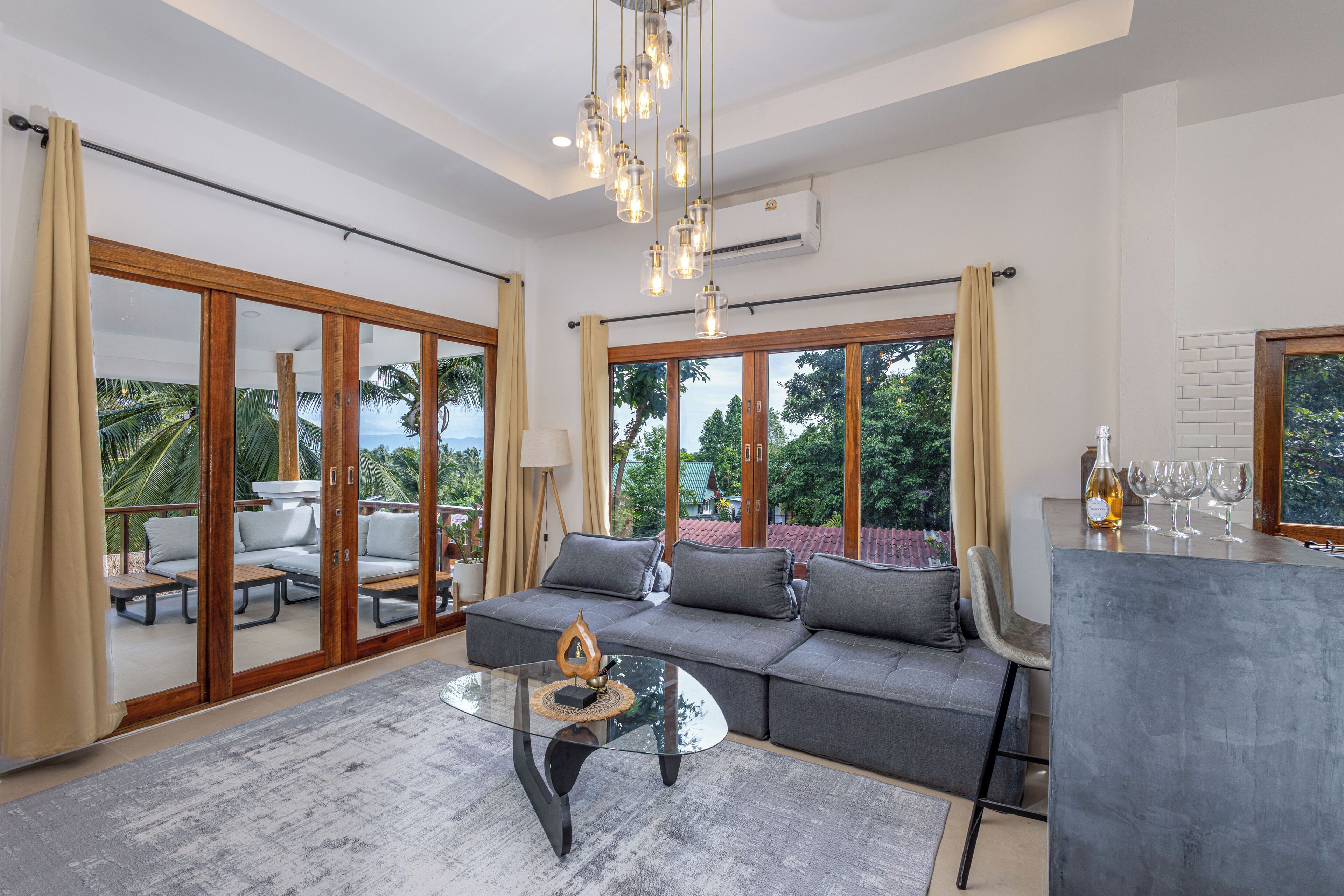 A spacious living area with a grey sofa, glass coffee table, and a bar area with a bottle and glasses. Large windows offer a view of palm trees and a red-roofed building.