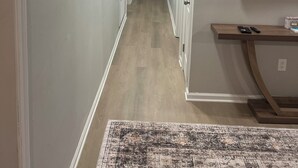 A hallway with a wooden floor, a patterned rug, a flat-screen TV mounted on the wall, and a table with remote controls.