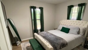 A bedroom with a large bed, a bench, two windows with green curtains, a ceiling light, and a door.