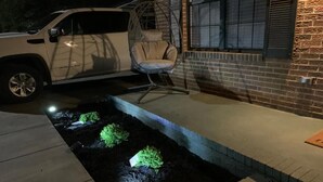 A driveway with a landscaped garden bed, a hanging chair, and a brick building in the background.