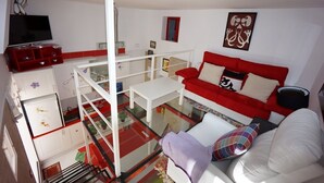 Un ambiente compatto con un divano rosso, un divano bianco e un tavolino in vetro. Una TV è montata a parete e ci sono diversi oggetti sugli scaffali.