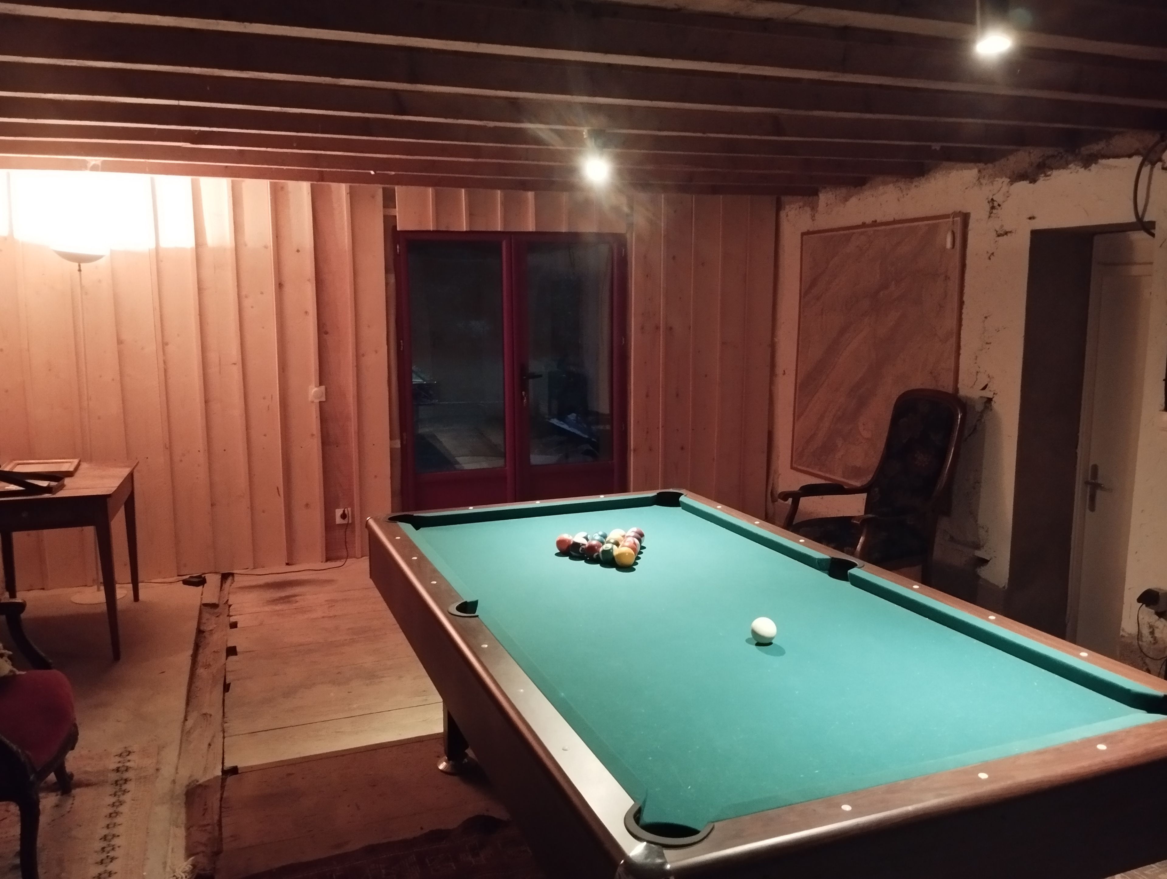 A pool table with balls, a chair, and a table in a room with wooden walls and a door.
