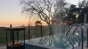Una piscina con borde de baldosas, un hogar para fuego y vistas a árboles y al atardecer.