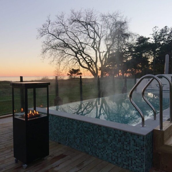 A swimming pool with a tiled edge, a fire pit, and a view of trees and a sunset.