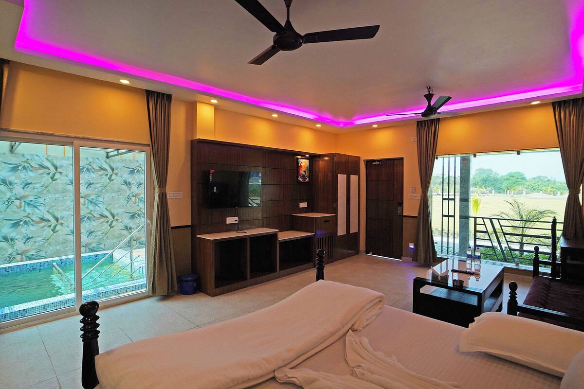 A hotel room with a bed, a TV, a ceiling fan, and a balcony with a view of the pool.
