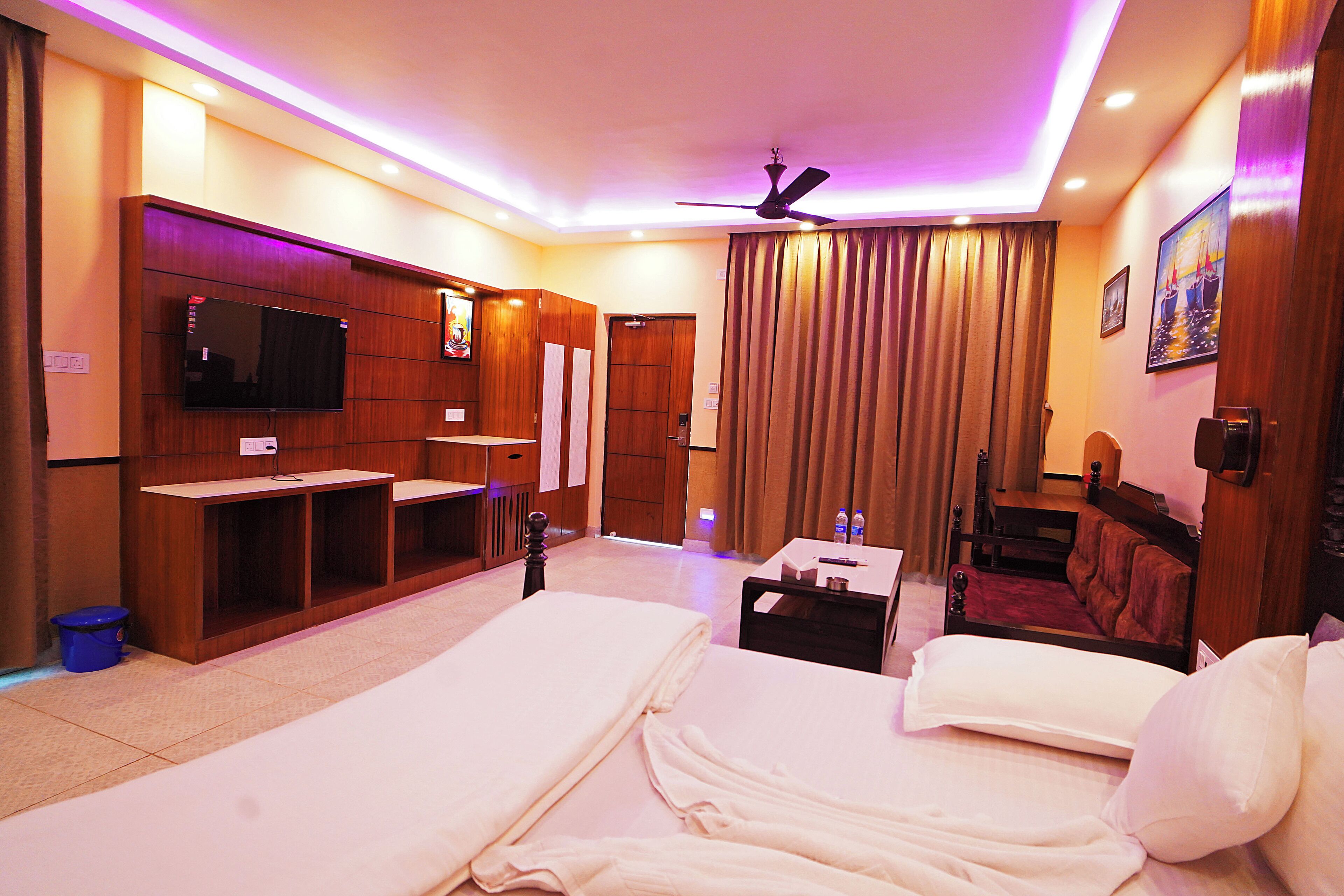 Habitación de hotel con dos camas, televisión, escritorio, ventilador de techo y un ventanal grande con cortinas.