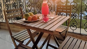 Een houten tafel met een roze drankje, fruit en een glas op een balkon met uitzicht op een zonsondergang.