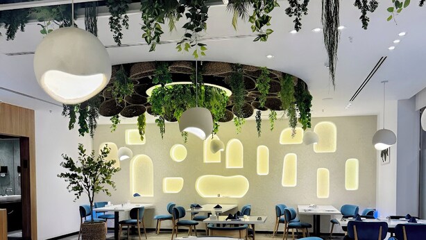 Een moderne ruimte met ronde tafels en blauwe stoelen, met een plafond vol hangende planten en verlichte wandpanelen.