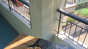 Een eettafel met een bord eten, een glas en bestek op een balkon met uitzicht op gebouwen en een straat.