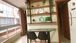 A small kitchen with a glass table and two chairs, a refrigerator, and a window with a view of a building. - SaltBeach Atalaia Residence Inn - N102 (Salinópolis)