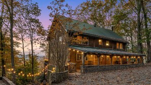 Une cabane rustique avec des fondations en pierre, un revêtement extérieur en bois et un toit végétalisé. Elle est dotée de nombreuses fenêtres et d’un porche illuminé de guirlandes lumineuses.