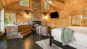 Une chambre située dans un chalet en rondins, avec une cheminée en pierre, un grand lit, un lavabo en pierre et un téléviseur fixé au mur.