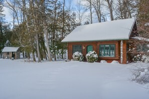 A wooden cabin with a snow-covered roof, surrounded by trees and a snowy landscape.