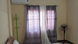 Une chambre à coucher avec un lit, des rideaux, un climatiseur, un ventilateur sur pied et une plante verte.