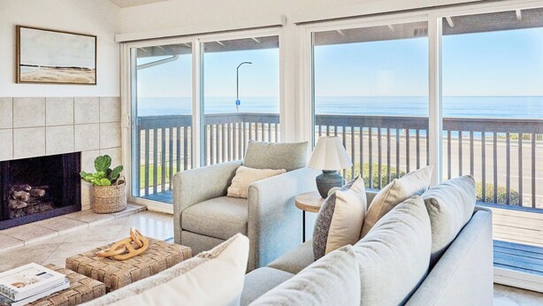 A living room with a fireplace, a sofa, a chair, and a view of the ocean through large windows.