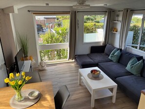 A modern living room with a sofa, a dining table with a vase of flowers, and a view of a garden through large windows. - RBR 511 - Beach Resort Kamperland (Kamperland)