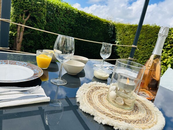 Une table dressée à l’extérieur, avec une bouteille de champagne, des verres, un saladier rempli de fruits et un set de table en fibres tressées.