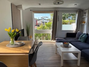 A modern living room with a sofa, a dining table with chairs, and a view of the outdoors through large windows.