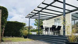 A covered outdoor area with a wooden deck, a seating arrangement, and a white building with a glass door.