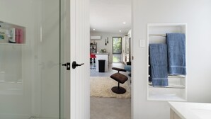 A modern bathroom with a glass shower enclosure, a white vanity with a towel rack, and a visible kitchen area in the background.