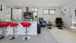 A modern kitchen with white cabinets, a stainless steel refrigerator, and a bar area with red stools. There are two chairs and a small table with a vase of flowers.
