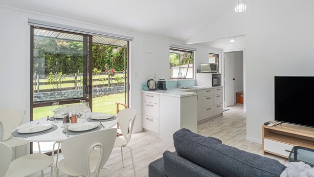 A modern kitchen with white cabinets, a dining area with a table set for four, and a grey sofa in the living area. - Rewa Rewa Retreat - Tairua Holiday Home (Tairua)