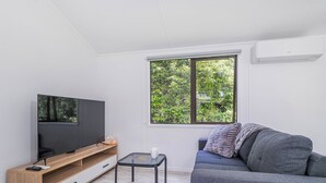 A modern living room with a flat-screen TV on a wooden TV stand, a gray sofa with a blanket, a small table with a cup, and a window with a view of greenery. - Rewa Rewa Retreat - Tairua Holiday Home (Tairua)