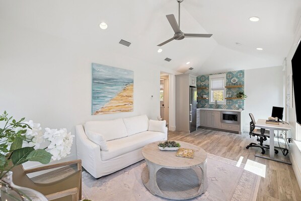 Un salon avec un canapé blanc, une table basse ronde, un ventilateur de plafond et un tableau représentant une plage.
