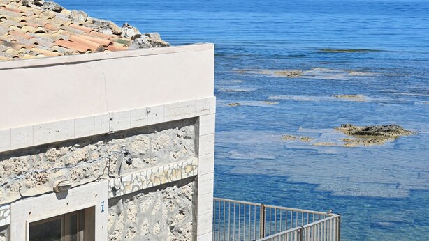 Ένα παραθαλάσσιο κτίριο με πρόσοψη από πέτρα, ένα μπαλκόνι με μεταλλική μπαλκονόπορτα και ανεμπόδιστη θέα στη θάλασσα.