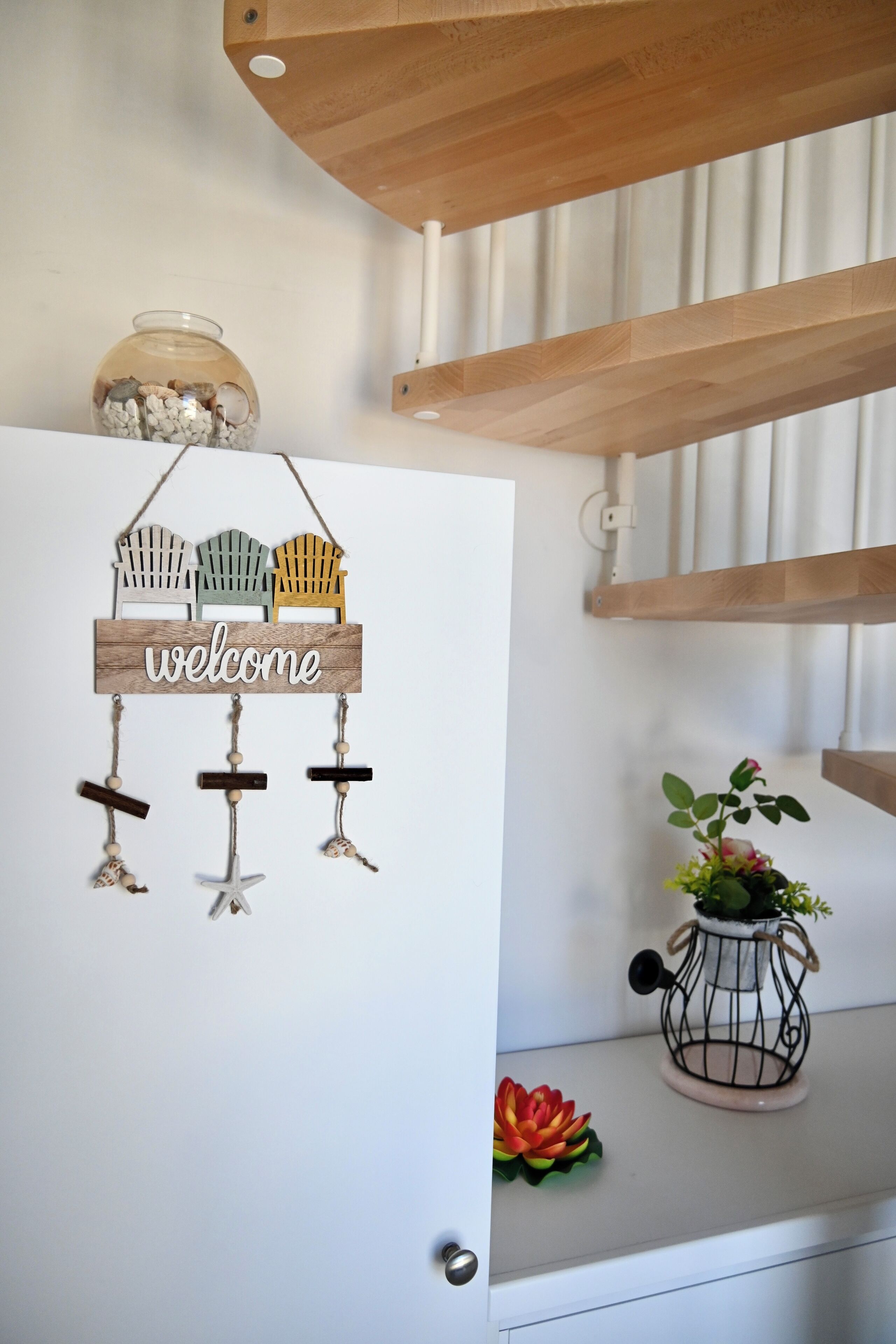 A welcome sign with wooden Adirondack chairs and a glass jar with pebbles on a white door.