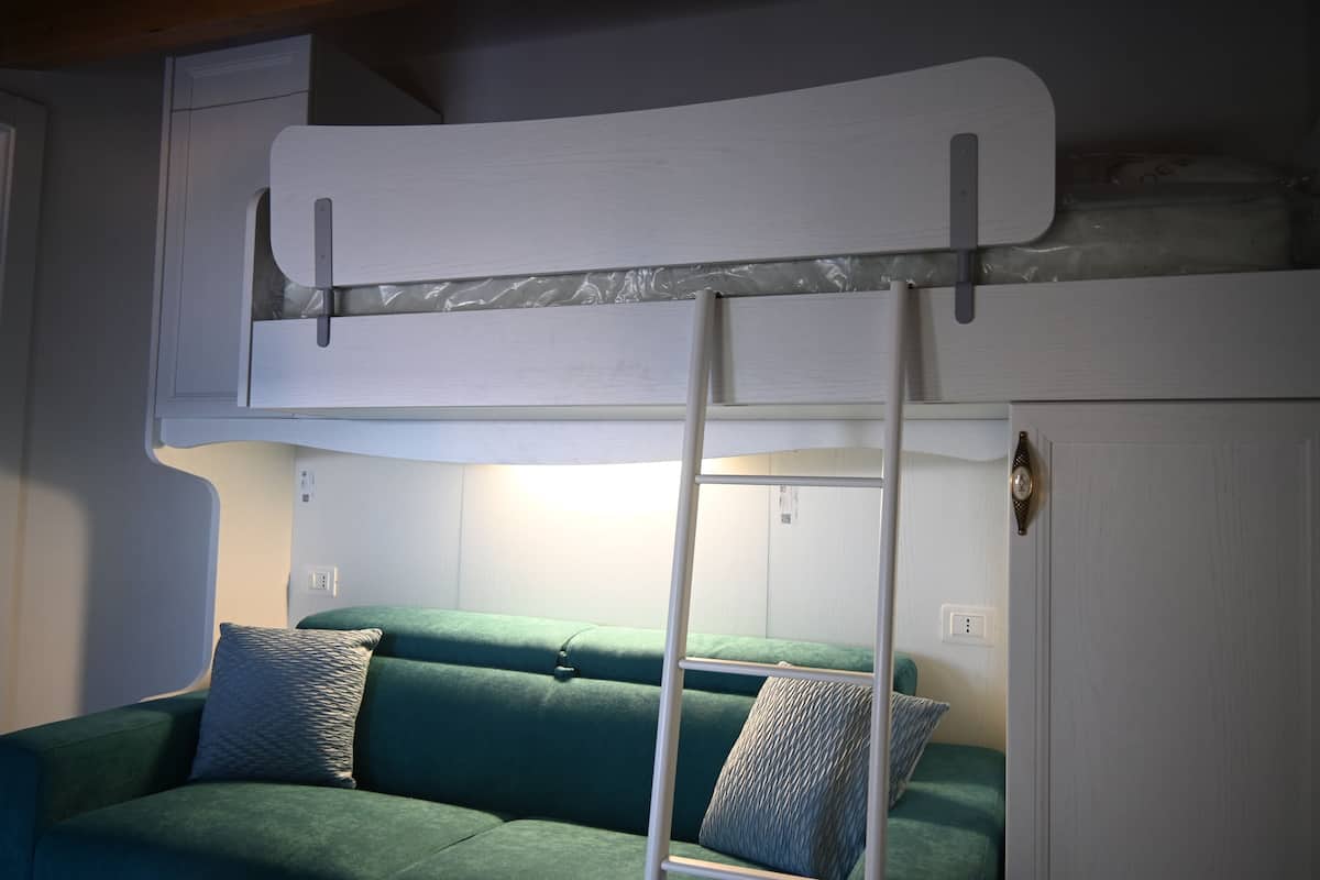 Ein Stockbett mit einer Leiter, ein grünes Sofa mit gemusterten Kissen und eine weiße Wand mit Lichtschalter.