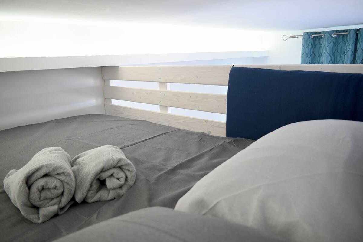 Ein Einzelbett mit grauem Matratze und blauer Decke, ein Holz-Kopfteil und ein Fenster mit Vorhängen.