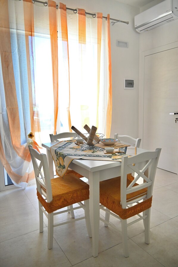 Un comedor con una mesa y sillas blancas, un mantel estampado y una ventana con cortinas translúcidas.