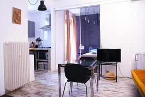 A compact living space with a kitchenette, a dining area with a table and chairs, a bed with a TV, and a window with curtains.