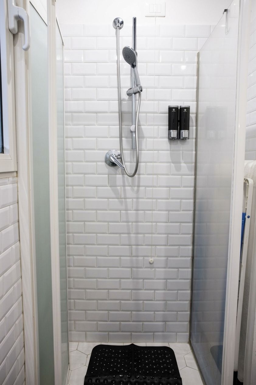 Een kleine, afgesloten douchecel met witte betegelde wanden en een zwarte mat op de vloer.