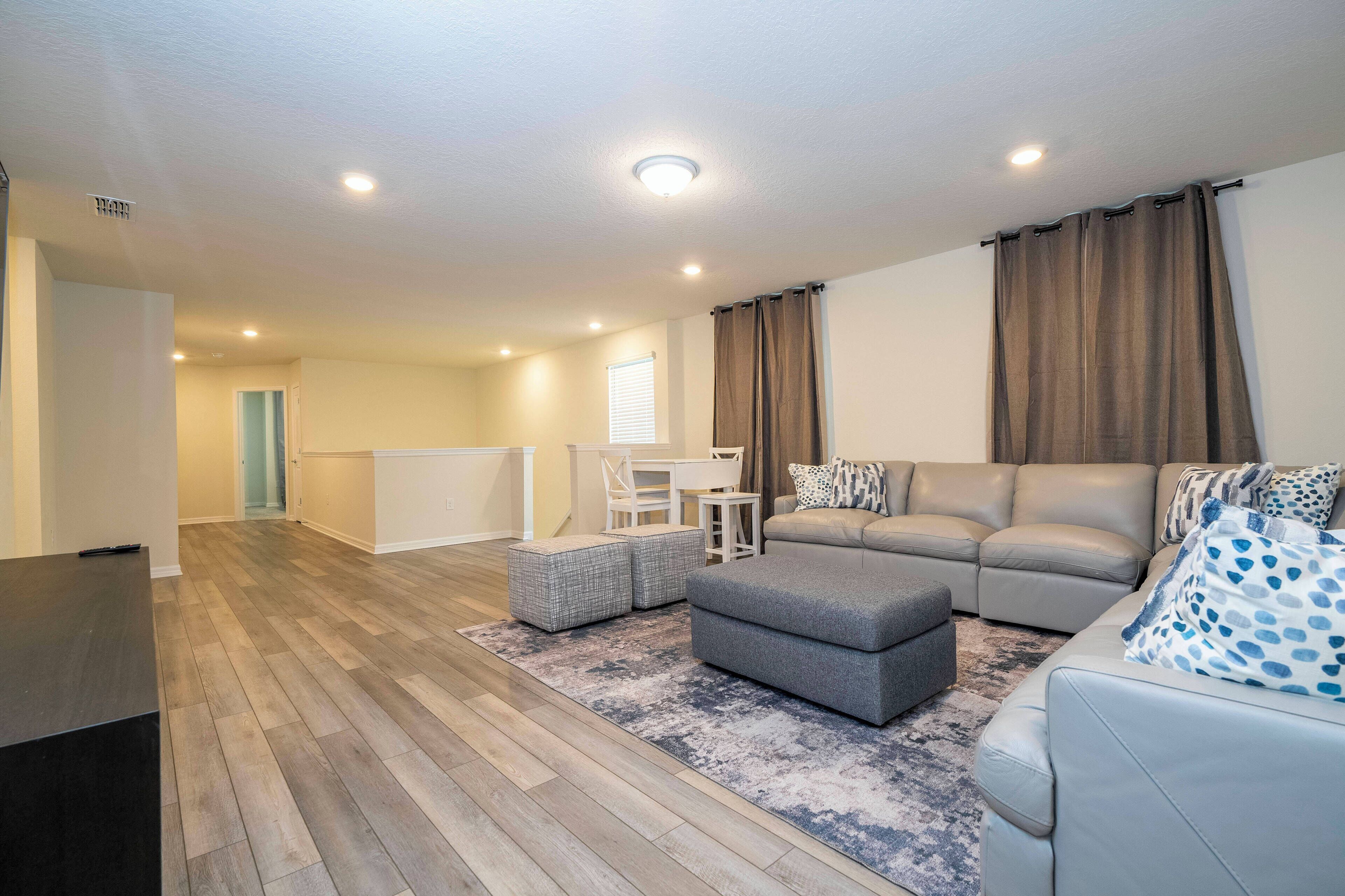 Ein modernes Wohnzimmer mit grauem Sofa, Hocker und einem Essbereich mit Tisch und Stühlen.