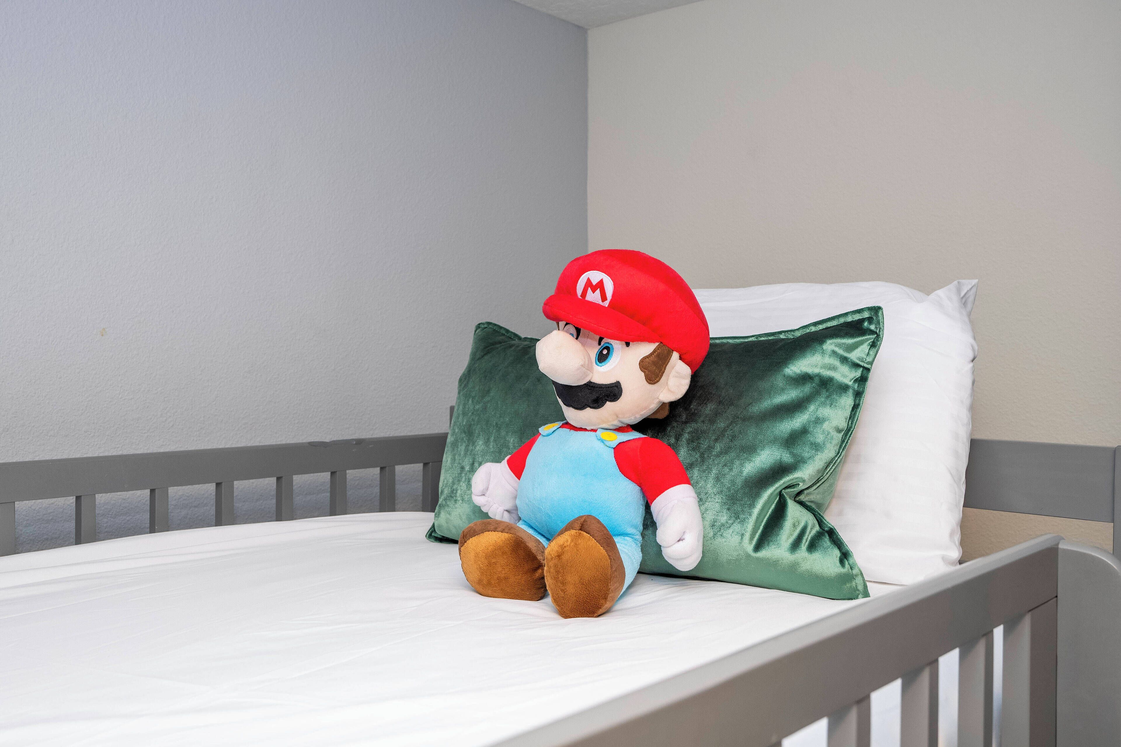 A plush Mario toy sitting on a bed with a green pillow and white sheets.