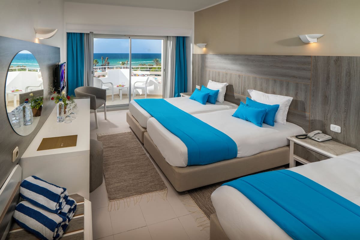 A hotel room with two beds, a desk with a mirror, a TV, and a balcony with a view of the sea.