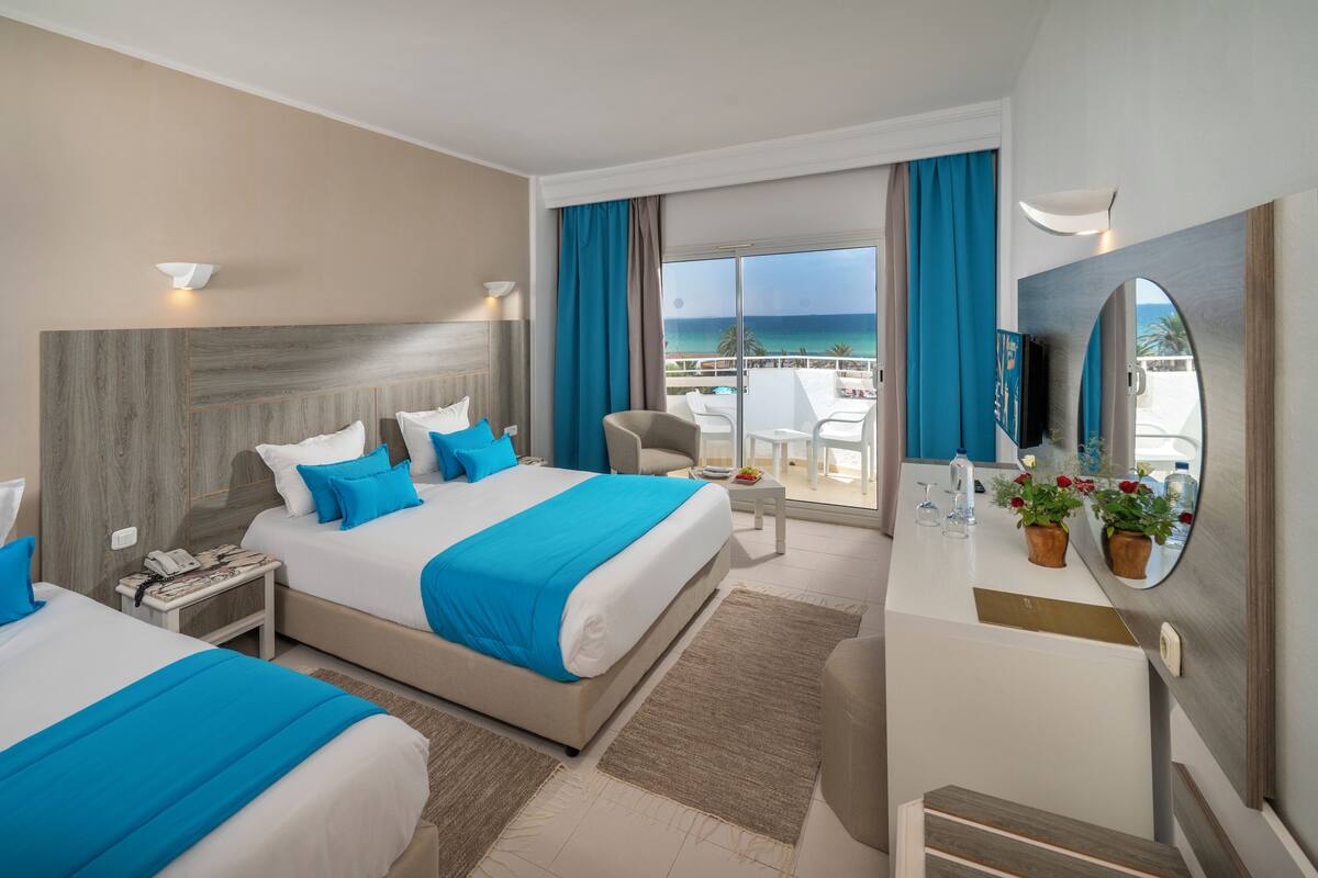 A hotel room with two beds, a desk, a chair, a balcony with a view of the sea, and a large mirror.