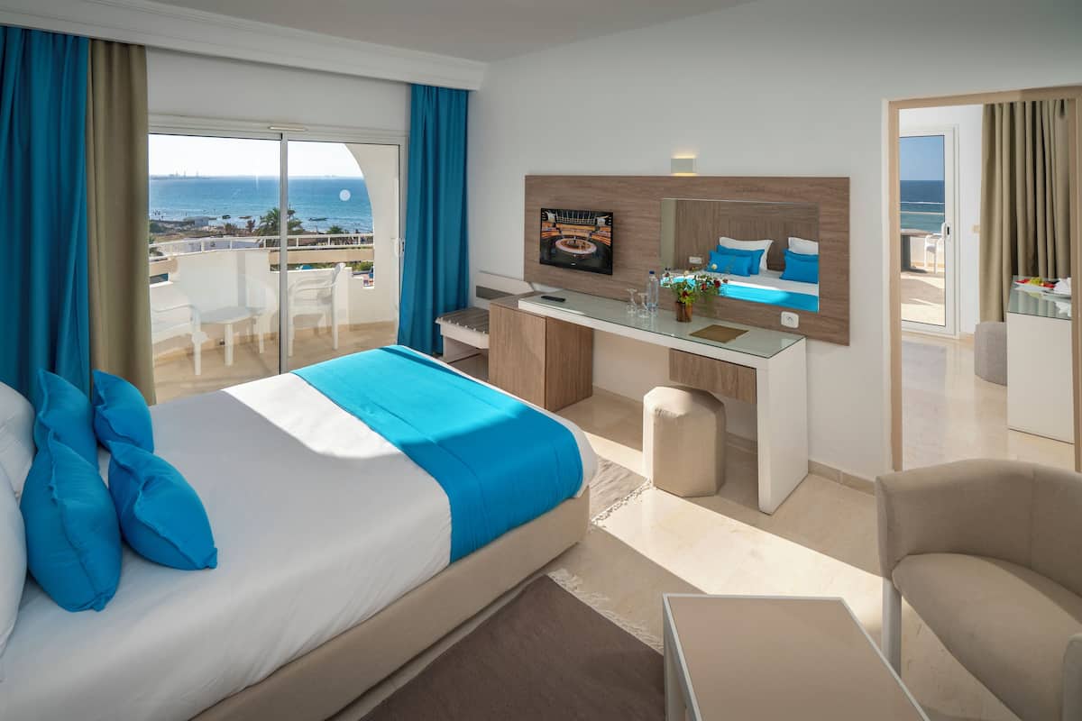 A modern hotel room with a large bed, a desk, a TV, and a balcony with a view of the sea.