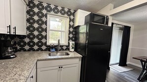 A kitchen with white cabinets, a black refrigerator, a coffee maker, and a window with a view of trees. - Rayben Residence - Centrally Located (Jacksonville)