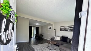 A modern living room with a dark wood floor, a grey sofa, a coffee table, and a dining area with a table and chairs. - Rayben Residence - Centrally Located (Jacksonville)