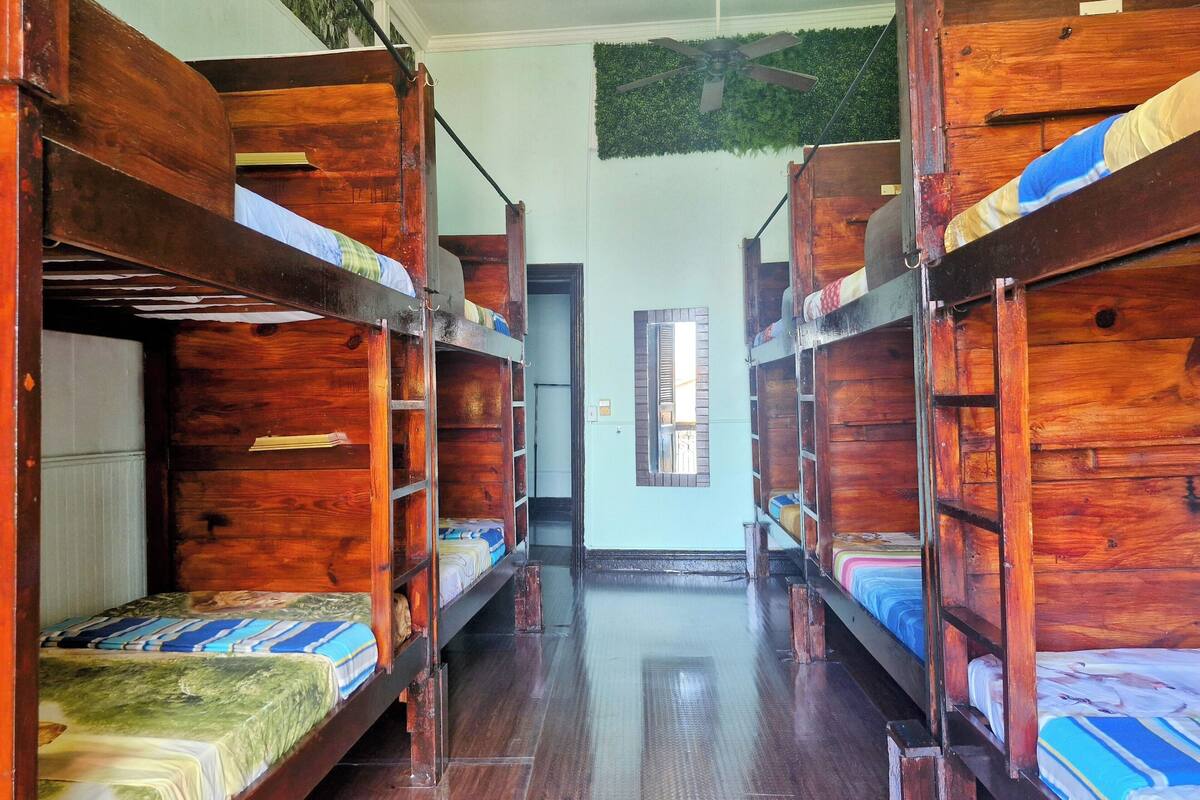 Un dormitorio con literas de madera, suelo de madera, ventilador de techo y ventana con vistas al exterior.