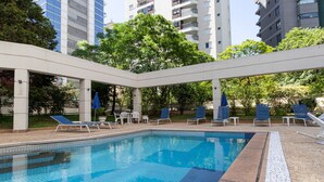 A swimming pool with lounge chairs and umbrellas, surrounded by greenery and high-rise buildings. - Elegance Suítes Vila Olímpia (Sao Paulo)