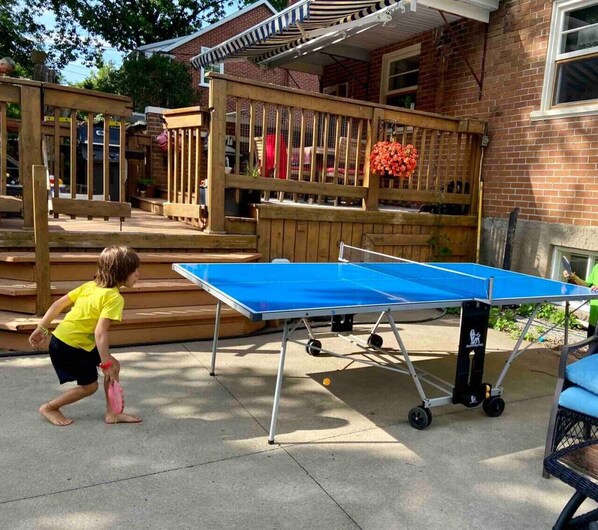 Une table de ping-pong dans un jardin, avec une terrasse en bois et une maison en briques en arrière-plan.