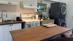 Une cuisine compacte avec des meubles blancs, un plan de travail en bois, une cuisinière, un four à micro-ondes, un lave-linge et une chaise.